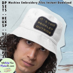 Może przedstawiać: Biały kapelusz typu bucket z czarną naszywką, na której widnieje napis "It's too peopley outside" w żółtym kolorze. Kapelusz nosi osoba o kręconych brązowych włosach.