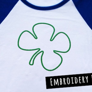 Puede incluir: Una camiseta blanca con un trébol de cuatro hojas bordado en verde. El texto "EMBROIDERY FILE" está impreso en negro en la parte inferior de la camiseta.