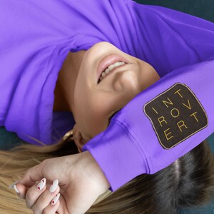 May include: A woman wearing a purple hoodie with a black patch that says "INTROVERT" in gold lettering.