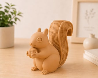 Puede incluir: Figura de ardilla de color marrón claro sosteniendo una nuez. La ardilla tiene una cola texturizada y está sentada. La figura está sobre una superficie de madera clara, con una planta y un marco de fotos en el fondo.