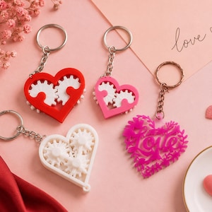 May include: Heart-shaped keyrings in red, pink, white, and fuchsia. The pendants feature gear designs and the word "love". The keyrings have silver and bronze-coloured rings.