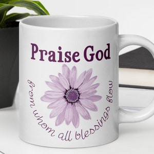 May include: White ceramic mug with the text "Praise God" in a dark purple font, above a light purple daisy illustration. The phrase "from whom all blessings flow" is written in a circular pattern around the flower.