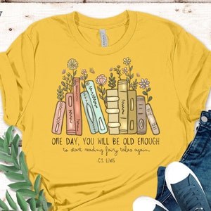 May include: A yellow t-shirt with a stack of books and flowers printed on the front. The text on the shirt reads "ONE DAY, YOU WILL BE OLD ENOUGH to start reading fairy tales again - C.S. LEWIS".