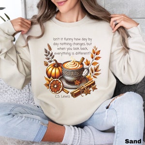 May include: A beige sweatshirt with a graphic design of a cup of coffee with whipped cream, a pumpkin, an orange slice, cinnamon sticks, and fall leaves. The text "Isn't it funny how day by day nothing changes, but when you look back, everything is different?" and "C.S. Lewis" are printed on the sweatshirt.