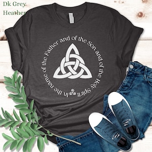 May include: Dark grey heather t-shirt with a white Celtic knot design and the text "In the name of the Father and of the Son and of the Holy Spirit." The shirt is paired with blue jeans and dark blue sneakers.