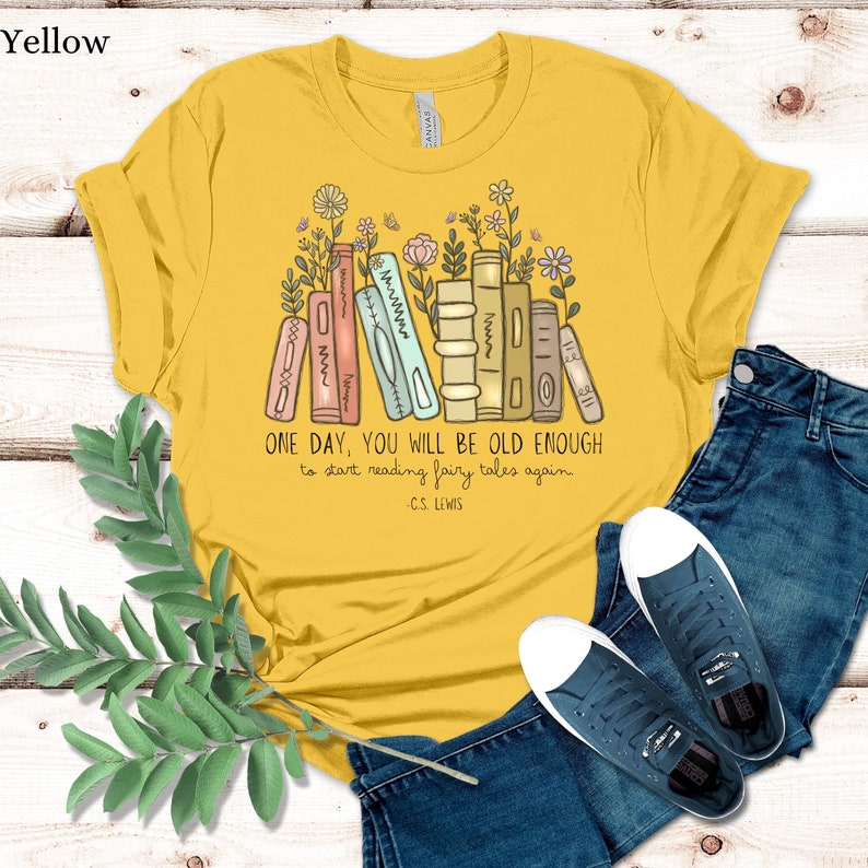 May include: A yellow t-shirt with a stack of books and flowers printed on the front. The text on the shirt reads "ONE DAY, YOU WILL BE OLD ENOUGH to start reading fairy tales again - C.S. LEWIS".