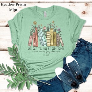 May include: A light green t-shirt with a stack of books and flowers. The text on the shirt reads "ONE DAY, YOU WILL BE OLD ENOUGH to start reading fairy tales again - C.S. LEWIS"