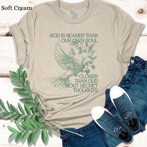 May include: A beige t-shirt with a green dove and leaves design. The text "God is nearer than our own soul" and "Closer than our most secret thoughts" is printed on the shirt.