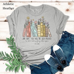 May include: A heather grey t-shirt with a graphic of a stack of colorful books with flowers growing from the top. The text "ONE DAY, YOU WILL BE OLD ENOUGH to start reading fairy tales again. -C.S. LEWIS" is printed below the graphic.