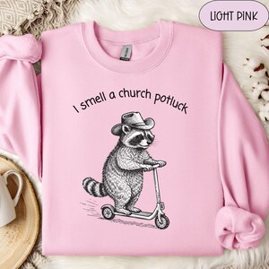 May include: Light pink sweatshirt with the text "I smell a church potluck." A black and white illustration of a raccoon wearing a cowboy hat and riding a scooter is printed on the front. The sweatshirt has long sleeves and a crew neck.