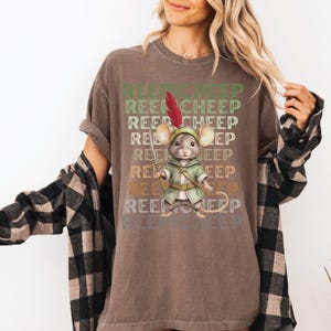 May include: A brown t-shirt with a vintage-style graphic of a mouse wearing a green tunic and holding a sword. The text "Reepicheep" is repeated in a faded, layered design.