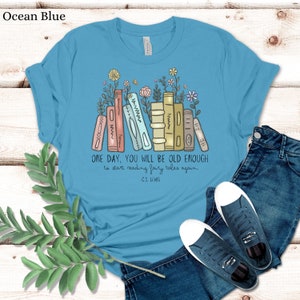 May include: A light blue t-shirt with a graphic of a stack of books with flowers growing from the top. The text "ONE DAY, YOU WILL BE OLD ENOUGH to start reading fairy tales again - C.S. LEWIS" is printed below the graphic.