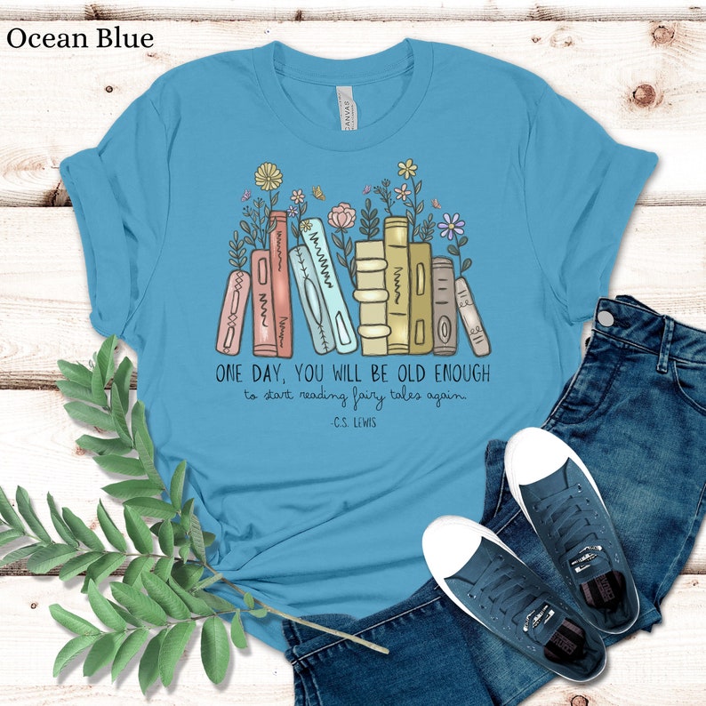 May include: A light blue t-shirt with a graphic of a stack of books with flowers growing from the top. The text "ONE DAY, YOU WILL BE OLD ENOUGH to start reading fairy tales again - C.S. LEWIS" is printed below the graphic.