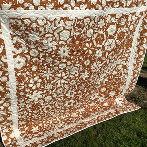 May include: A brown and white quilt with a floral pattern. The quilt is hanging on a clothesline and has a white border.