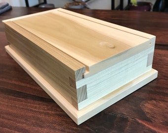 May include: A wooden box with a lid, crafted from light wood with a natural finish. The box features a simple rectangular design and a smooth surface. The lid is slightly raised and has a subtle groove running along the top. The box is resting on a dark wooden surface.