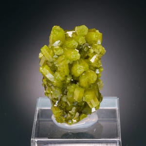 Può includere: Un gruppo di cristalli verde lime vibranti, possibilmente un campione minerale, è esposto su un supporto trasparente e rettangolare. I cristalli hanno una struttura geometrica e angolare, con una superficie lucida e riflettente. Lo sfondo è una sfumatura di grigio scuro.