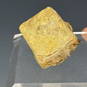 May include: A close-up of a small, square, yellow-brown mineral specimen. The surface has a rough, textured appearance with visible cracks and variations in color. The mineral is displayed on a clear, angled surface against a dark background.