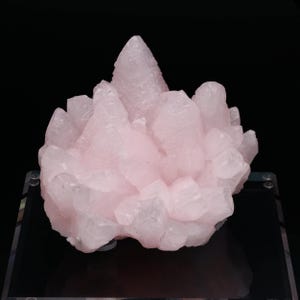 Peut inclure: Un amas de cristaux de quartz rose pâle, à structure cristalline. Les cristaux sont disposés en formation groupée, avec un pic central proéminent. Le fond noir met en valeur la couleur et la texture du minéral.