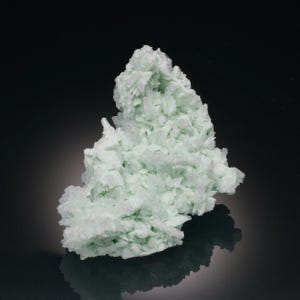 May include: A light green mineral specimen with a crystalline structure. The mineral has a clustered, layered appearance, with a slightly rough texture. The specimen is set against a dark background, with a reflection visible below.