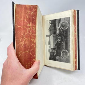 Op de afbeelding: Een open antiek boek met een zwarte rug en rood en goud gemarmerde schutbladen. Op de rechterpagina is een zwart-witfoto van een vintage auto te zien. Het boek wordt door een hand opengehouden.