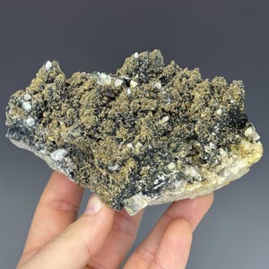Pode incluir: Um espécime mineral natural áspero com uma superfície texturizada. A rocha apresenta uma mistura de formações cristalinas cinza escuro, bege e brancas. O mineral é segurado em uma mão contra um fundo cinza.