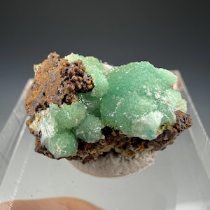 May include: A mineral specimen featuring clusters of vibrant mint green, crystalline formations attached to a dark, textured, rust-coloured rocky matrix. The green crystals exhibit a sparkling, druzy-like surface texture with visible radial patterns.