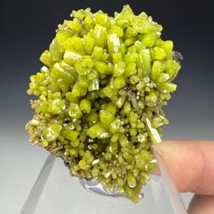Puede incluir: Un vibrante espécimen mineral de color verde lima. La estructura cristalina presenta numerosos cristales prismáticos, densamente empaquetados. El color del mineral es un verde brillante, casi neón, con un aspecto ligeramente translúcido. La base es marrón oscuro y terroso.
