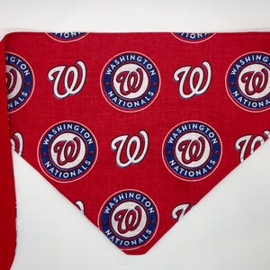 Può includere: Bandana rossa con un logo bianco e blu dei Washington Nationals ripetuto su tutto il tessuto. La bandana ha una cravatta rossa su un lato.
