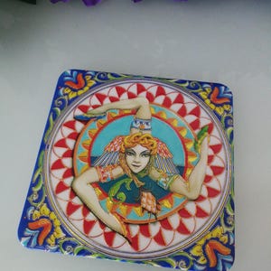 May include: Square ceramic tile featuring a detailed illustration of a female figure with wings, a crown, and a trident. The design is set against a backdrop of red and white patterns, with a blue and gold border. The tile is likely a decorative item.
