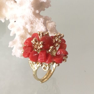 May include: A gold ring with a red coral flower design. The ring is made of small pieces of coral that are arranged in a flower shape. The ring is set on a white background.