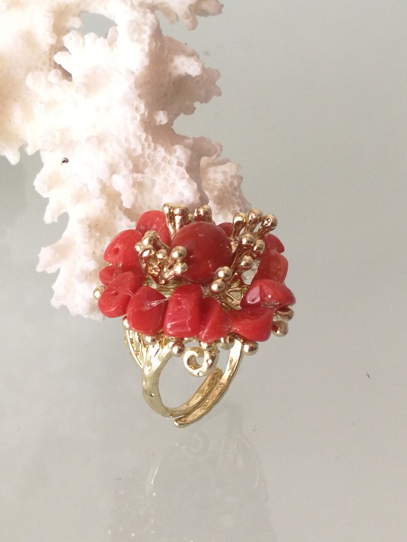 May include: A gold ring with a red coral flower design. The ring is made of small pieces of coral that are arranged in a flower shape. The ring is set on a white background.