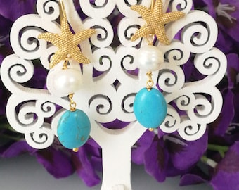 Puede incluir: Un par de pendientes colgantes con un dije de estrella de mar en tono dorado en la parte superior, seguido de una perla blanca redonda y terminado con una piedra turquesa ovalada. Los pendientes cuelgan de ganchos de metal dorado y tienen textura.