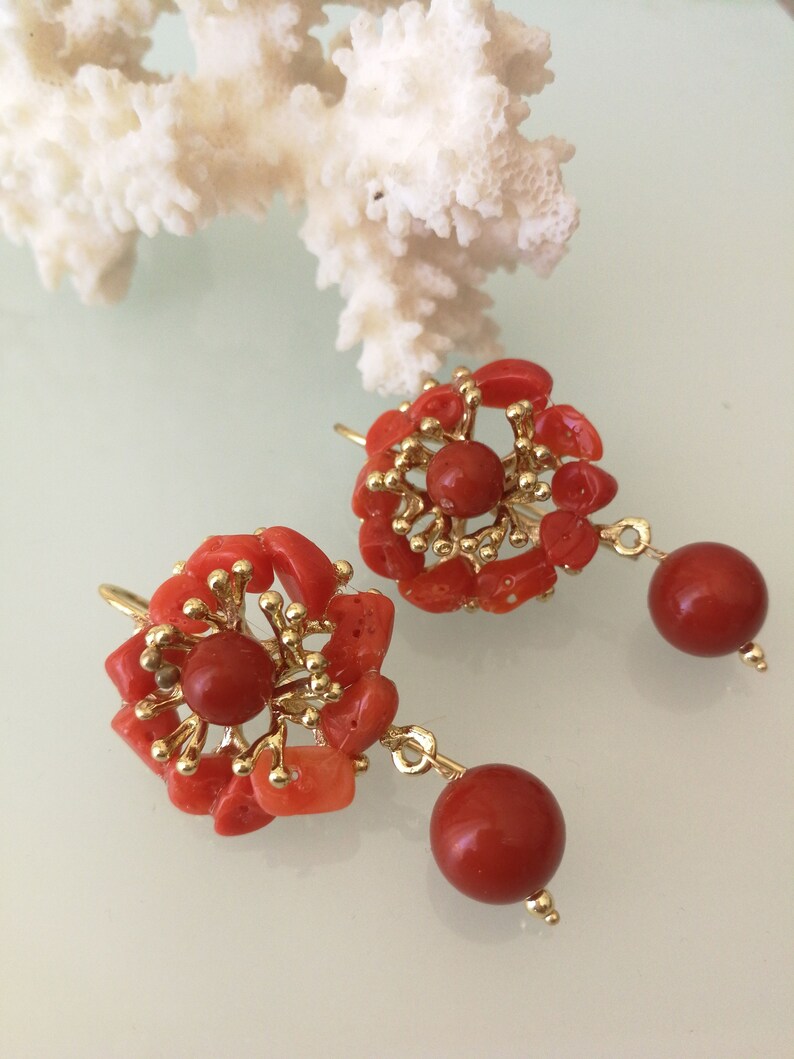 May include: A pair of coral earrings with gold accents. The earrings are made of small pieces of coral that are arranged in a flower shape. The earrings have a drop design with a round coral bead hanging from the flower.