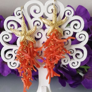 May include: A pair of gold starfish earrings with dangling coral branches. The coral is a vibrant orange color and the starfish are detailed with a textured surface.