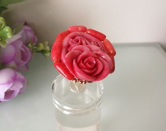 May include: A ring featuring a large, carved pink rose centerpiece surrounded by a halo of smooth, cylindrical red coral beads. The floral design is detailed with layered petals, and the band is constructed from small, metallic gold-toned beads.