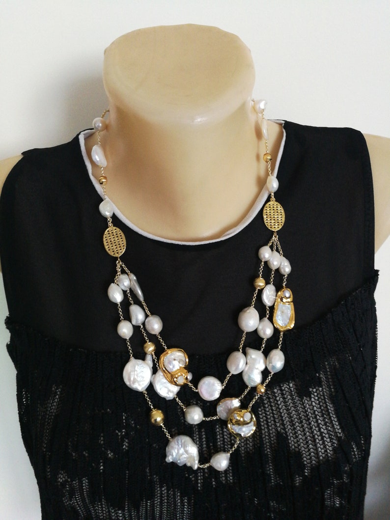 May include: A multi-strand necklace with large white pearls and gold accents. The necklace features a variety of pearl shapes, including round and oval. The gold accents include small beads and larger, decorative pieces.