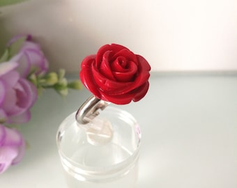 May include: A ring featuring a bright red, carved rose-shaped centerpiece mounted on a polished silver-toned metal band. The rose design displays detailed, layered petals that create a realistic floral appearance, securely attached to the metallic ring base.