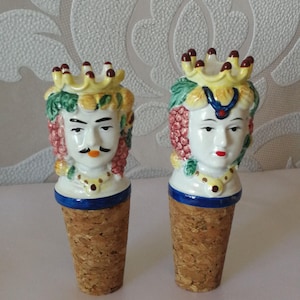 May include: Two decorative ceramic bottle stoppers. Each stopper features a detailed face with a crown, mustache, and grape-like hair. The stoppers have a cork base and are painted with vibrant colours, including yellow, red, and green.