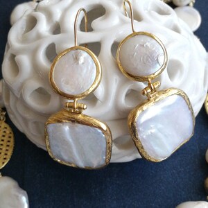 May include: A pair of gold earrings with white pearl-like drops. The earrings have a square and round shape. The earrings are on a white surface.