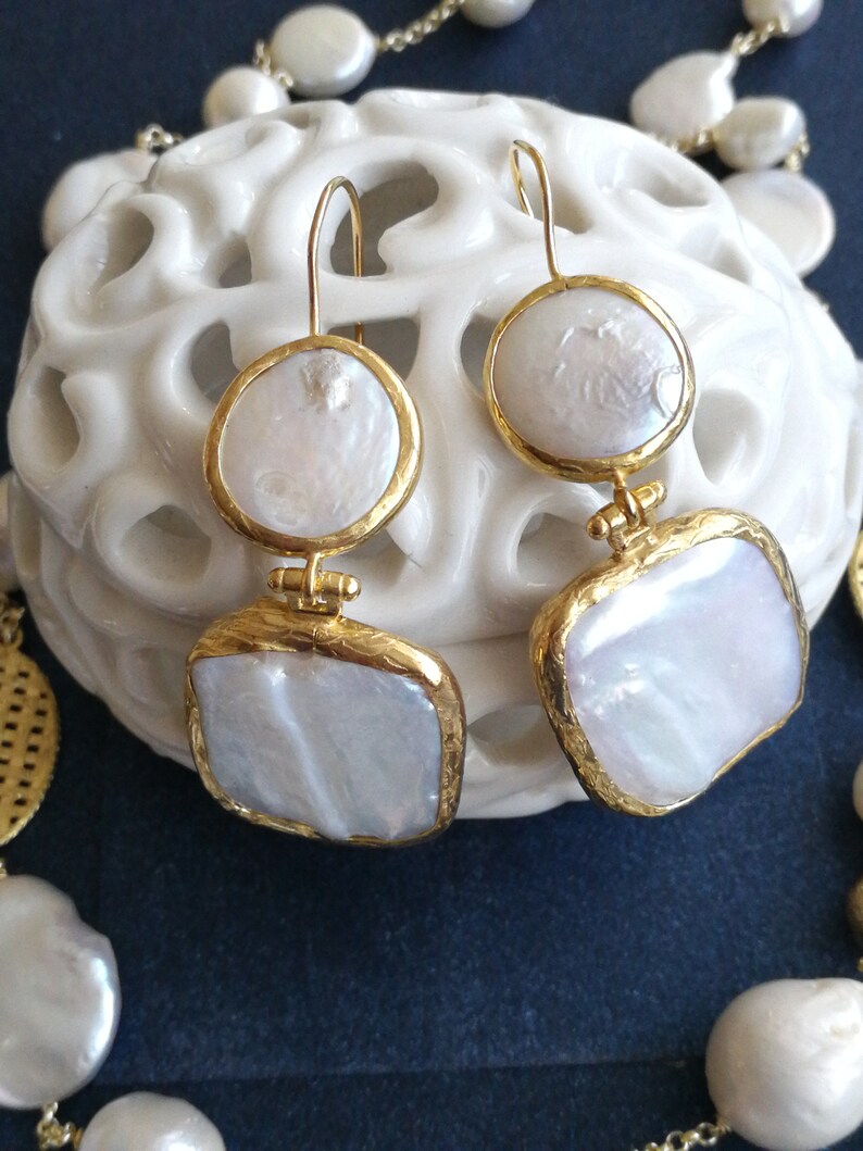 May include: A pair of gold earrings with white pearl-like drops. The earrings have a square and round shape. The earrings are on a white surface.