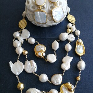May include: A gold chain necklace with white baroque pearls and gold accents. The necklace is layered and features large, irregular shaped pearls. The necklace is paired with matching gold earrings with white baroque pearls.