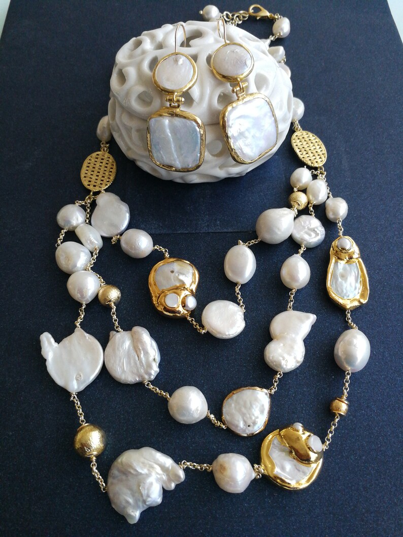May include: A gold chain necklace with white baroque pearls and gold accents. The necklace is layered and features large, irregular shaped pearls. The necklace is paired with matching gold earrings with white baroque pearls.