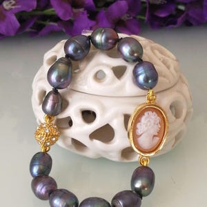 Peut inclure: Un bracelet en perles noires avec un charme en or représentant un camée de profil de femme. Le bracelet est sur un fond blanc.