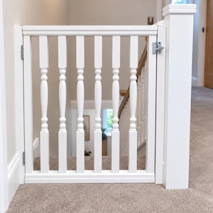 May include: A white wooden safety gate installed at the top of a staircase. The gate features vertical balusters with a turned design, a horizontal top and bottom rail, and a silver metal latch mechanism attached to a matching white post.