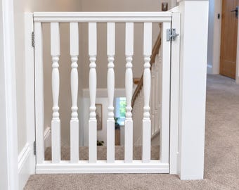 May include: A white wooden safety gate installed at the top of a staircase. The gate features vertical balusters with a turned design, a horizontal top and bottom rail, and a silver metal latch mechanism attached to a matching white post.