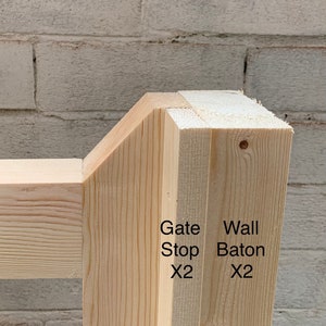 May include: Close-up of a wooden gate stop and wall baton, both made of unfinished wood. The gate stop is labelled "Gate Stop x2" and the wall baton is labelled "Wall Baton x2".