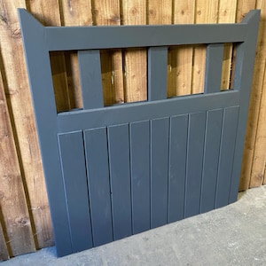 May include: A dark grey wooden gate with a slatted design and a decorative top. The gate has a horizontal row of rectangular openings in the top section.