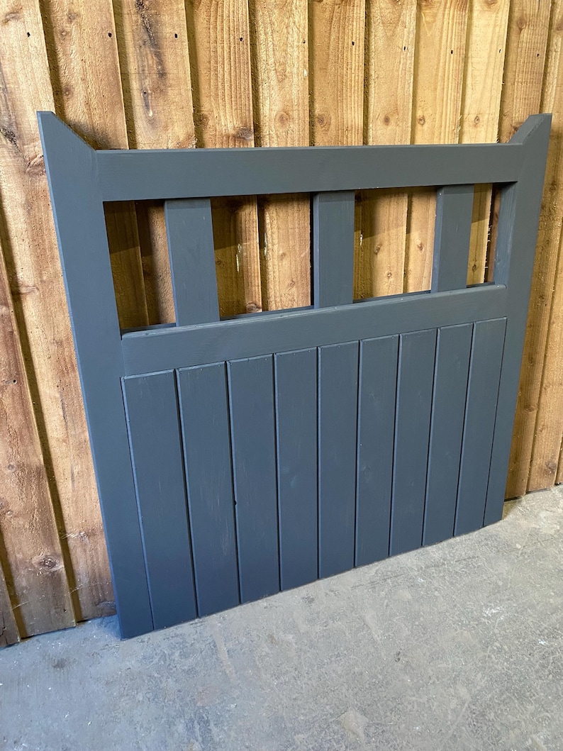May include: A dark grey wooden gate with a slatted design and a decorative top. The gate has a horizontal row of rectangular openings in the top section.