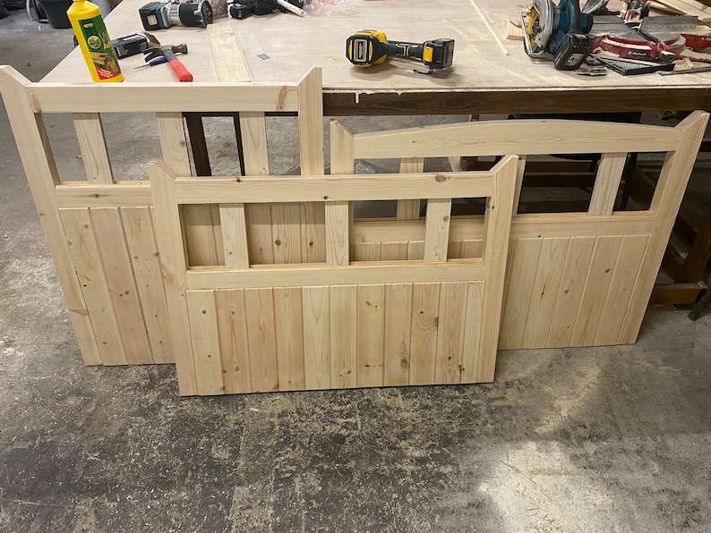 May include: Three unfinished wooden gate panels with a slatted design and a decorative top rail. The panels are arranged on a light wood surface.
