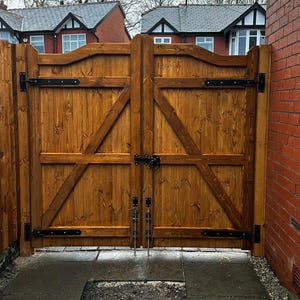 May include: A pair of wooden double gates with black metal hinges and latches. The gates are closed and are in front of a brick wall.